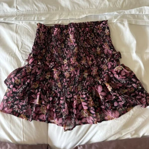 Blommig volangkjol från Neo Noir - Supersöt kort kjol från Neo Noir med blommigt mönster i rosa, lila och beige på svart botten. Kjolen har smockad midja och flera volanger som ger en flowy känsla. Perfekt för dig som gillar romantisk och trendig stil.