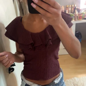 Vinröd volangtopp - Säljer en helt ny vinröd topp med volangdetalj runt halsringningen. Toppen är figurnära och har en strukturerad, lätt skrynklig yta. Perfekt för dig som vill ha något enkelt men ändå med en twist.