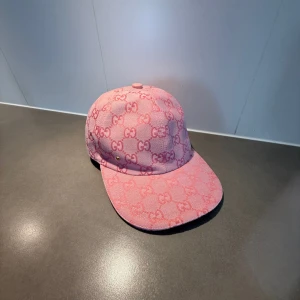 Rosa Gucci keps - Säljer en rosa keps från Gucci med klassiskt GG-monogram i hela tyget. Kepsen har böjd skärm och justerbart spänne bak i metall. Materialet är kraftig bomullscanvas och insidan är ljus. Perfekt för dig som gillar lyxiga accessoarer.