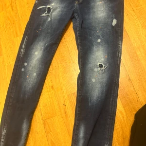 Blå slitna jeans med hål - Säljer ett par blå jeans med slitningar och hål på flera ställen. Jeansen har en smal passform och coolt tvättad look med ljusa partier och färgstänk. Perfekta för dig som gillar en trendig och avslappnad stil.