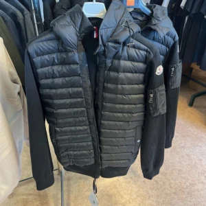 Svart vadderad hoodie från Moncler - Säljer en svart vadderad hoodie från Moncler med quiltade paneler framtill och huva. Jackan har långärmade ärmar i mjukt material och en diskret Moncler-logga på ärmen. Perfekt för dig som vill ha en stilren och sportig look.