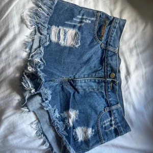 Blå jeansshorts med fransar och slitningar - Säljer ett par blå jeansshorts från Wrangler med råa fransar i bensluten och slitna detaljer både fram och bak. Klassisk femficksmodell med knapp och dragkedja. Perfekta för sommaren!
