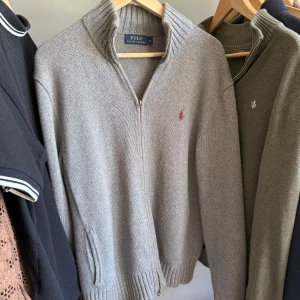 Grå stickad tröja med dragkedja från Polo Ralph Lauren - Säljer en klassisk grå stickad tröja från Polo Ralph Lauren med dragkedja och hög krage. Tröjan har ribbade muddar och en liten broderad logga på bröstet. Perfekt för lager-på-lager och stilren look.