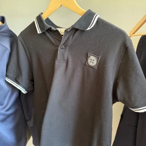 Svart piké från Stone Island - Säljer en svart pikétröja från Stone Island med vit rand på krage och ärmslut. Tröjan har korta ärmar, knappslå framtill och klassisk logotyp på bröstet. Slim fit-modell som passar perfekt till en stilren look.