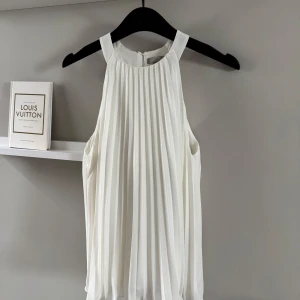 Vit plisserad ärmlös blus - Superfin vit blus från H&M med plisserat tyg och hög hals. Blusen är ärmlös och har en luftig, A-linjeformad passform som ger ett elegant intryck. Perfekt för sommarens tillfällen och enkel att styla med accessoarer.