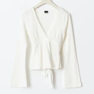 Vit omlottopp med knytband - Säljer en vit omlottopp med v-ringning och knytband i midjan. Toppen har långa ärmar och är tillverkad i ett lätt, strukturerat material. Perfekt för lager-på-lager eller som en enkel bas i garderoben. 