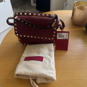 Valentino Vinröd axelväska med nitar från  - Säljer en vinröd axelväska från Valentino med guldfärgade nitar längs kanterna och axelremmen. Väskan har ett stilrent, fyrkantigt format och är tillverkad i skinn. Invändigt finns flera fack och den stängs med magnetlås. Perfekt accessoar för att lyfta din outfit.