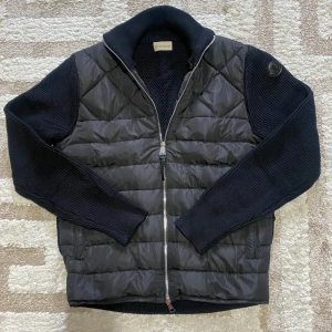 Moncler cardigan - Snygg svart hybridjacka från Moncler med quiltad framsida i syntet och stickade ärmar och rygg. Jackan har dragkedja framtill och hög krage. Moncler-logga på ärmen ger en exklusiv touch.