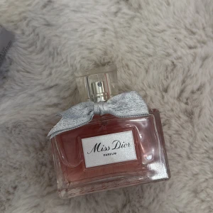 Miss Dior Parfum - Säljer en elegant parfym från Dior, Miss Dior. Flaskan är rektangulär i glas med rosa doftvätska och dekorerad med en söt silverfärgad rosett vid halsen. Perfekt för dig som gillar lyxiga och feminina dofter.