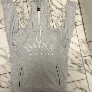 Grå hoodie från Hugo Boss - Säljer en ljusgrå hoodie från Hugo Boss med stor vit logga på bröstet och dragsko i huvan. Tröjan har fickor och är tillverkad i mjukt material. Perfekt för en avslappnad stil. Storlek S perfekt skick