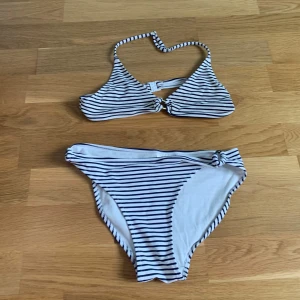 Randig bikini från H&M - Säljer en vit bikini med smala svarta ränder från H&M. Överdelen har halterneck med knyt i nacken och spänne bak, och bikinitrosan har en liten knutdetalj på sidan. Perfekt för sommaren!