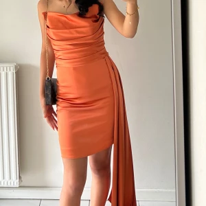 Orange axelbandslös klänning i satin - Säljer en snygg orange axelbandslös klänning i satin med draperad överdel och asymmetrisk kjol. Klänningen har en lång detalj på sidan som ger extra effekt. Perfekt för dig som vill sticka ut!Från Beauty queens, kan passa både 32 och 36