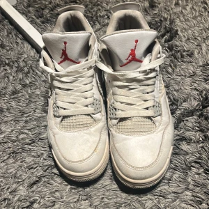 Vita Nike Air Jordans 4 Retro - Säljer ett par vita Nike Air Jordans 4 Retro med röda detaljer på plösen och gråmelerad sula. Skorna har klassisk snörning, meshpaneler och den ikoniska Jumpman-loggan på hälen. Perfekta för dig som gillar streetwear och sneakers med retrovibbar. Kom med prisförslag 