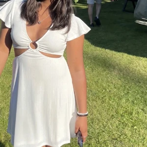 Vit kortklänning med ringdetalj från Hm - Säljer en söt vit kortklänning från hm med v-ringning och en snygg ringdetalj framtill. Klänningen har korta ärmar och lätt utställd kjol. Perfekt till sommaren. Använda 2 gånger 