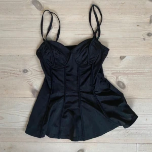 Svart korsett-topp från H&M Divided - Säljer en svart korsett-topp från H&M Divided i storlek XXS. Toppen har smala axelband, markerade sömmar och lätt utsvängd nederdel. Materialet är mjukt och glansigt, vilket ger en snygg look. kan tyvärr inte skicka bild på då den inte passar💕💕