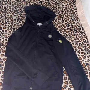 Svart hoodie från Stone Island - Säljer en svart hoodie från Stone Island med dragkedja och huva. Klassisk logotyp-patch på ärmen och två fickor framtill. Perfekt för dig som gillar streetwear och vill ha något stilrent med ikonisk design.