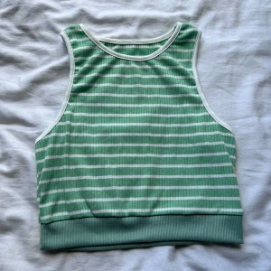 Grön och vit randig ribbad linnetopp - Snygg ribbad linnetopp i grönt och vitt med horisontella ränder och vita kanter. Toppen är ärmlös och har en bred mudd nedtill för en sportig look. Perfekt för varma dagar eller som ett lager under en skjorta.