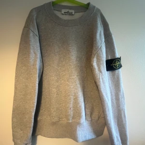 Grå sweatshirt från Stone Island - Säljer en klassisk grå sweatshirt från Stone Island med rund hals och den ikoniska loggan på vänster ärm. Tröjan är i mjuk bomull och har ribbade muddar vid ärmslut och nederkant. Perfekt för en avslappnad stil.