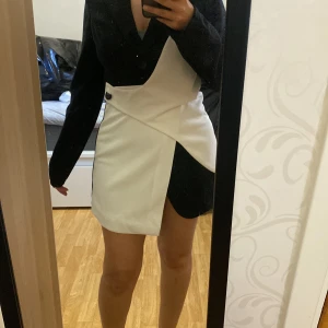 Svart och vit omlottklänning - Säljer en snygg svart och vit kortklänning med omlottdetalj framtill. Klänningen har lång ärm och en cool kontrast mellan svart och vitt tyg. Perfekt för dig som vill sticka ut med en modern look.