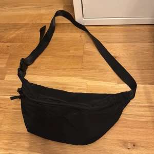 Svart magväska från Uniqlo - Säljer en stilren svart magväska från Uniqlo. Väskan har justerbart band och dragkedja upptill. Perfekt för att bära med sig det viktigaste och har flera fack inuti för bättre organisering. Enkel och snygg design som passar till många tillfällen.