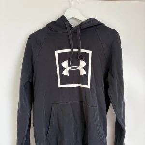 Svart hoodie från Under Armour - Säljer en svart hoodie från Under Armour med stor vit logga på bröstet. Tröjan har huva med dragsko och en stor magficka. Perfekt för träning eller chill. Klassisk och enkel design.