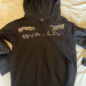 Svart hoodie med strasstenar från Bvaldi - Svart hoodie från Bvaldi i storlek XS med dragkedja och huva. Framsidan har coola motiv och text i strasstenar som ger extra bling. Perfekt för dig som gillar streetstyle och vill sticka ut lite extra. Mjuk insida och klassisk passform.