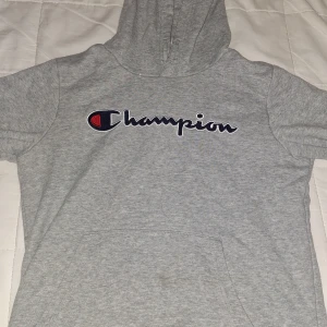 Grå hoodie från Champion med logga - Säljer en grå hoodie från Champion med stor broderad logga på bröstet. Tröjan har korta ärmar, huva och en klassisk känguruficka framtill. Perfekt för chill dagar eller när du vill ha en sportig look.