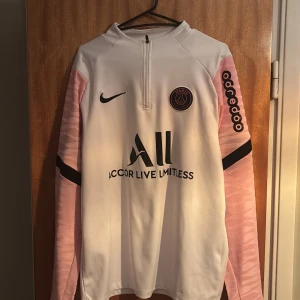 PSG långärmad träningströja Nike - Säljer en snygg långärmad Paris Saint-Germain träningströja från Nike. Tröjan är vit med ljusrosa ärmar och svarta detaljer, PSG-logga på bröstet och sponsortryck. Materialet är mjukt och sportigt, perfekt för fotbollsträning.