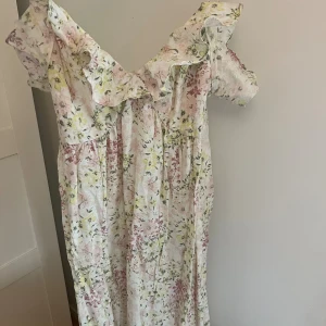 Blommig långklänning med volang - Superfin långklänning med blommigt mönster i ljusa toner av rosa, gul och grön på vit botten. Klänningen har volangdetalj längs urringningen och korta ärmar. Perfekt för sommarens alla tillfällen!