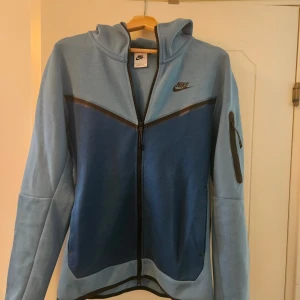 Blå Nike Tech fleece stl S - Nike hoodie i två nyanser av blått med hel dragkedja framtill och svart Nike-logga på bröstet. Tröjan har huva, lång ärm och en praktisk ficka med dragkedja på ena ärmen. Storlek S, men lite stor i storleken. Använd ett fåtal gånger, i mycket bra skick. 