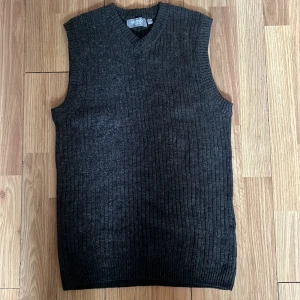 Mörkgrå stickad väst med v-ringning - Snygg mörkgrå stickad väst från M.DISO med v-ringad hals och ribbad struktur. Perfekt att slänga över en skjorta eller t-shirt för en chill men ändå uppklädd look. Klassisk passform och mjukt material som känns skönt mot huden.