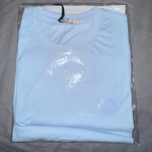 Ljusblå t-shirt från Moncler - Snygg ljusblå t-shirt från Moncler med rund hals och diskret Moncler-logga ton-i-ton på bröstet. T-shirten har en clean look och är gjord i mjuk bomull som känns skön mot huden. Perfekt för dig som gillar stilrena plagg med exklusiv känsla.