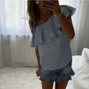 Ljusblå omlottop med volang - Supersöt ljusblå topp med omlottkänsla och volangdetalj över axeln. Toppen är oneshoulder och har en luftig, lätt struktur i tyget som känns som polyester. Perfekt för dig som vill ha en feminin och trendig look. Den har en liten defekt vid volangen