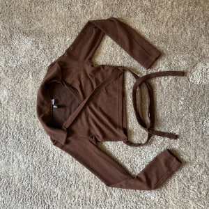 Brun omlottopp från H&M Divided - Superfin brun omlottopp från H&M Divided i ribbad bomull. Croppad modell med långa ärmar och knytband i midjan som ger en snygg siluett. Perfekt att styla med höga jeans eller kjol för en chill och trendig look.