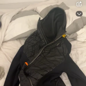 Svart hybridjacka från Parajumpers - Säljer en svart hybridjacka från Parajumpers med quiltade partier framtill och på ärmarna, samt mjuka sidor och huva. Jackan har dragkedja, ikonisk gul detalj vid halsen och Parajumpers-logga på ärmen. Perfekt för dig som gillar stilren och sportig look. Storlek L den är äkta har kvitto och påse 