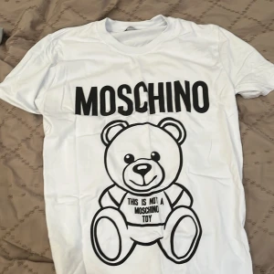 Vit Moschino t-shirt med björntryck - Vit t-shirt från Moschino med stort svart tryck av en söt björn och texten 'This is not a Moschino toy' på framsidan. Klassisk rund hals och korta ärmar. Tillverkad i mjuk 100% bomull, perfekt för dig som gillar statement-plagg och coola designer.