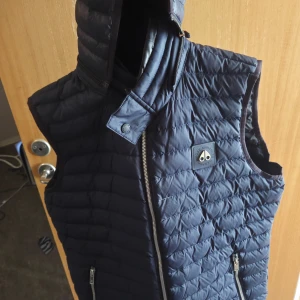 Moose knuckles väst - Snygg mörkblå dunväst från Moose Knuckles med quiltad design och hög krage. Västen har dragkedja framtill, två fickor med dragkedja och en ikonisk logotyp på bröstet. Perfekt lager-på-lager-plagg för kyliga dagar pris ej hugget i sten..