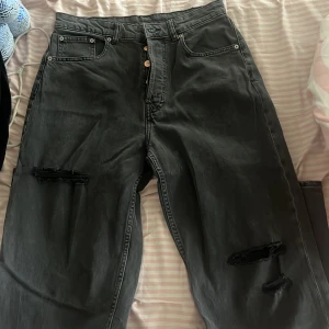 Svarta baggy jeans med slitningar - Säljer ett par svarta jeans med baggy passform och slitna detaljer på båda benen. Jeansen har klassisk femficksdesign, knäppning med knappar och är tillverkade i kraftigt denimtyg. Perfekta för en avslappnad och trendig look.