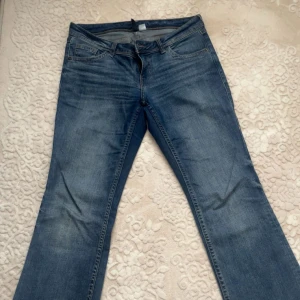 Blåa låg midjade bootcut jeans med fickdetaljer - Säljer ett par klassiska blå jeans med bootcut-modell och snygga fickor med knappar bak. Jeansen har en lätt tvättad look och vida ben, perfekt för dig som gillar retrostil. Materialet är jeans och färgen är en mellanblå ton.