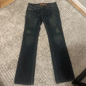 Mörkblåa jeans - Säljer ett par mörkblå jeans med klassiska kontrastsömmar och broderad logga på bakfickan. Jeansen här bootcut, slitna detaljer framtill och coola läderdetaljer vid midjan. Lite sönder längst midjan men inget som försämrar funktionen. För små för mig så finns ingen bild på
