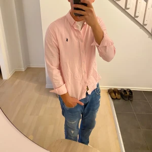 Ljusrosa skjorta från Ralph Lauren - Säljer en ljusrosa skjorta från Ralph Lauren med klassisk button down-krage och broderad logga på bröstet. Skjortan har lång ärm, normal passform och är tillverkad i mjuk bomull. Perfekt för dig som gillar stilrena och fräscha plagg.