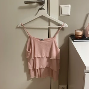Ljusrosa volanglinne från Kappahl - Supersöt ljusrosa linnetopp från Kappahl med tunna axelband och dubbla volanger. Toppen är ärmlös och har en flowy, luftig känsla som passar perfekt till varma dagar. Storlek 158/164