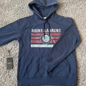 Marinblå Saint Laurent hoodie - Snygg marinblå hoodie från Saint Laurent med tryckt logga och ränder i rött och vitt på bröstet. Tröjan är i perfekt skick bytten är intressant!!! Pris kan diskuteras, hör av er för frågor