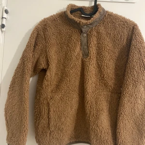 Brun teddy fleece-tröja med knappar - Supermjuk och fluffig brun teddytröja i fleece med hög krage och knappar framtill. Perfekt för kalla dagar, har långa ärmar och två fickor framtill. Oversized och cozy vibe, passar dig som gillar chill och avslappnad stil.