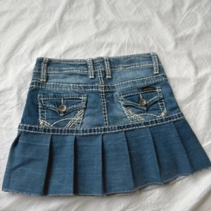 Jeanskjol från Cipo & Baxx, - Snygg blå jeanskjol från Cipo & Baxx med plisserad nederdel och coola bakfickor med dekorativa sömmar och knappar. Kjolen har bälteshällor och markerade kontrastsömmar för en extra detalj. Perfekt för dig som gillar street och denim vibes.