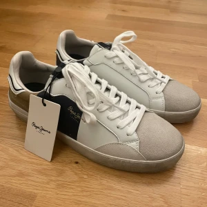 Pepe Jeans vita sneakers med mockadetaljer - Säljer nu mina HELT NYA Pepe jeans sneakers, då jag köpte fel storlek (nog dom skönaste sneakersarna jag någonsin prövat) Det är som sagt ett par snygga sneakers från Pepe Jeans i vitt skinn med grå mockatå, beige och mörkblå detaljer på sidor och häl. Klassisk låg modell med snörning och vit sula. Märkt med '1973' bak på hälen och Pepe Jeans-logga i guld på sidan. Perfekta för en stilren och trendig look Storlek: 42