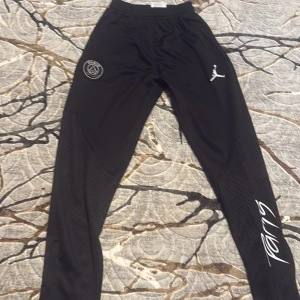 Svarta PSG x Jordan träningsbyxor - Snygga svarta träningsbyxor från Jordan x PSG med logga på höften och Jumpman-tryck på benet. Byxorna har elastisk midja, smal passform och coolt mönstrade paneler längs benen. Materialet är mjukt och lätt polyester, perfekt för sport eller chill.