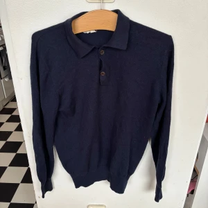 Mörkblå kashmirpiké från Soft Goat - Mörkblå långärmad pikétröja från Soft Goat i mjuk kashmir. Klassisk krage med två knappar framtill och ribbade muddar vid ärmslut och nederkant. Perfekt för dig som gillar stilren och bekväm look. Lappen där det står softgoat är lite trasig men annars bra skick.