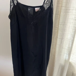 Svart spetsklänning från H&M - Svart klänning från H&M med tunna axelband och vacker spetsdetalj vid halsringningen. Klänningen har en rak och enkel siluett som ger en stilren look. Perfekt för dig som gillar minimalistisk stil med en touch av spets.