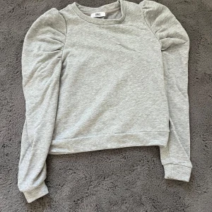 Grå sweatshirt med puffärmar från ONLY - Säljer en ljusgrå sweatshirt från ONLY med puffiga axlar och långa ärmar. Tröjan har en klassisk rund halsringning och är tillverkad i mjukt bomullsmaterial. Perfekt för dig som gillar en enkel men trendig look.
